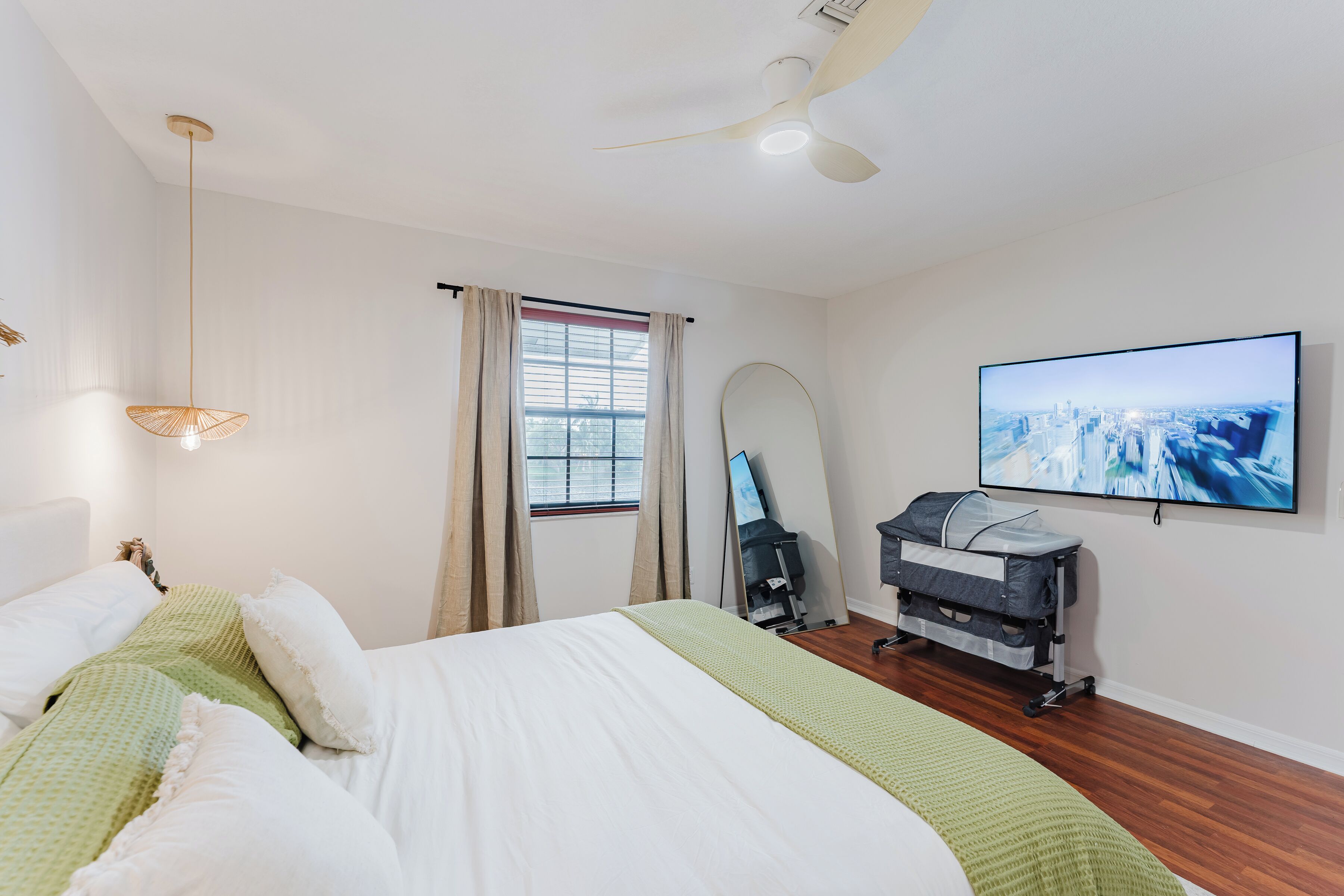 A hotel room with a bed, a ceiling fan, a TV mounted on the wall, and a window with curtains.