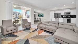 A modern living room with a beige sectional sofa, two armchairs, and a colorful geometric rug. A kitchen with white cabinets and stainless steel appliances is visible in the background.