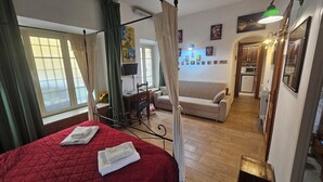 A bedroom with a red bedspread, a canopy bed, a sofa, and a kitchenette in the background. - Cavour 1&2 (Rome)