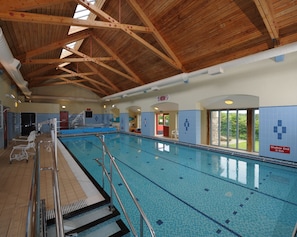 An indoor swimming pool with wooden beams, clear pool lanes, and a shallow end sign.