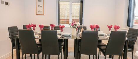 En spisestue med et rektangulært bord dækket til seks personer, omgivet af sorte stole. Bordet har blomsterarrangementer, og i baggrunden ses et vindue med persienner.