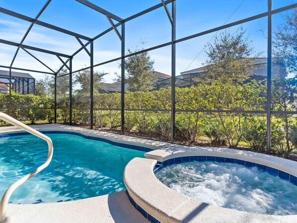 Une piscine avec un jacuzzi, entourée d’une structure en verre offrant une vue sur les arbres et les maisons.