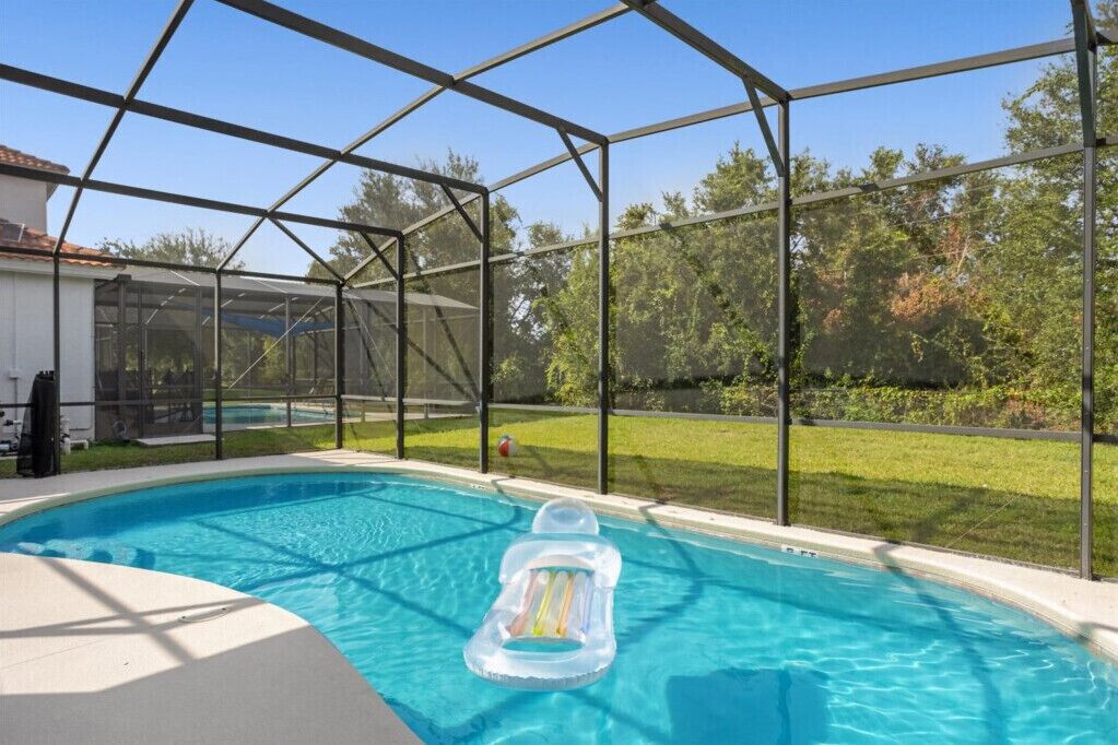 Piscina com boia inflável transparente, cercada por grade de metal, com gramado e árvores ao fundo.