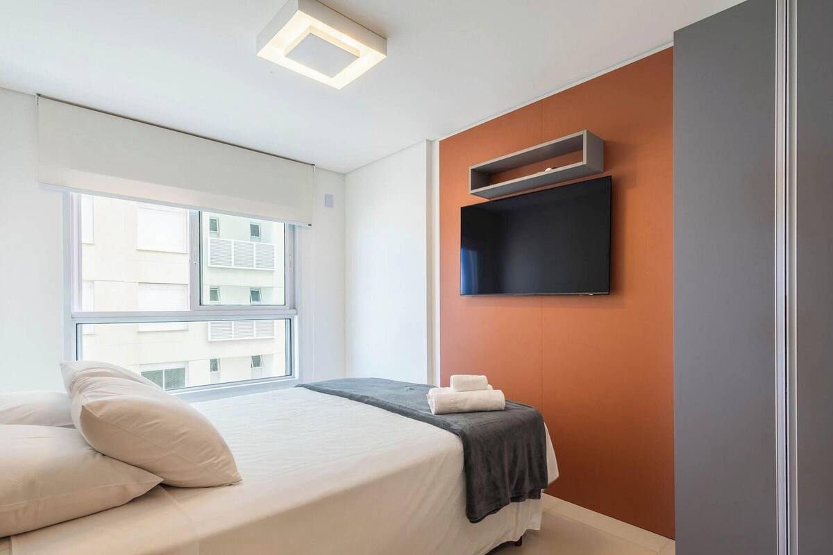 A modern hotel room with a large bed, a flat-screen TV mounted on the wall, and a window with a view of the building outside.