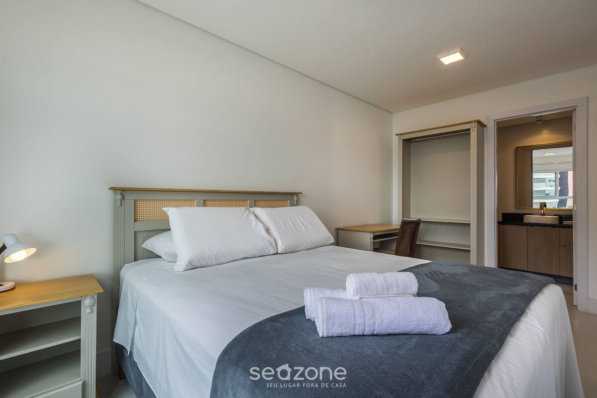 A neatly made bed with white linens and a grey blanket, a bedside table with a lamp, and a wooden nightstand.
