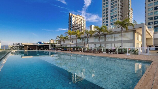 A swimming pool with lounge chairs and umbrellas, surrounded by tall buildings and palm trees. - Seafront Condominium With Pool - SHC (Picarras)