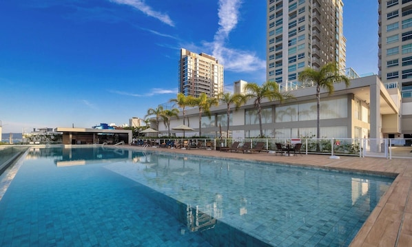 Una piscina con sillones de playa y sombrillas, rodeada de edificios altos y palmeras.