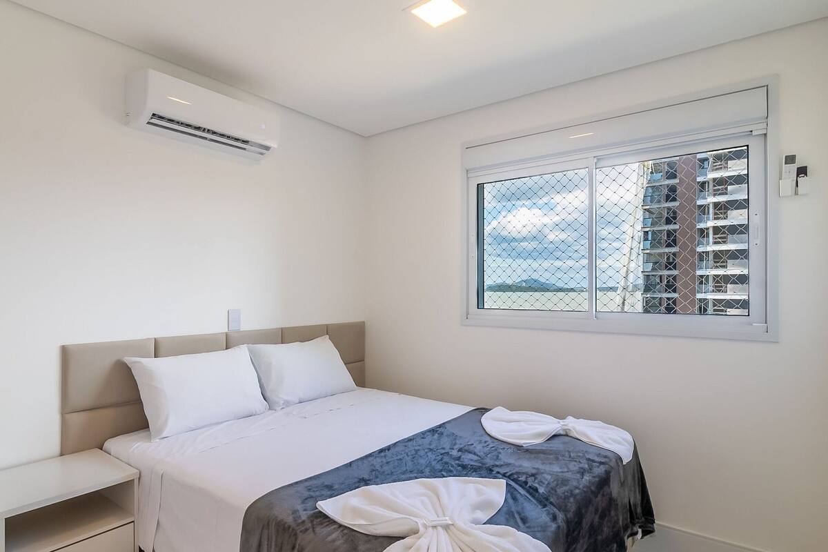 A bedroom with a bed, a nightstand, an air conditioning unit, and a window with a view of buildings.