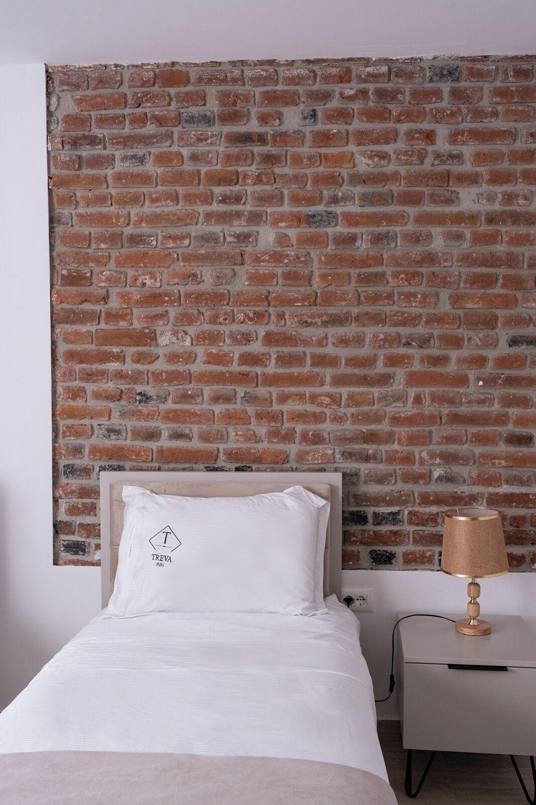Un dormitorio con cabecera de pared de ladrillo, cama con ropa de cama blanca, almohada con un logotipo, una lámpara de noche y una mesita de noche.