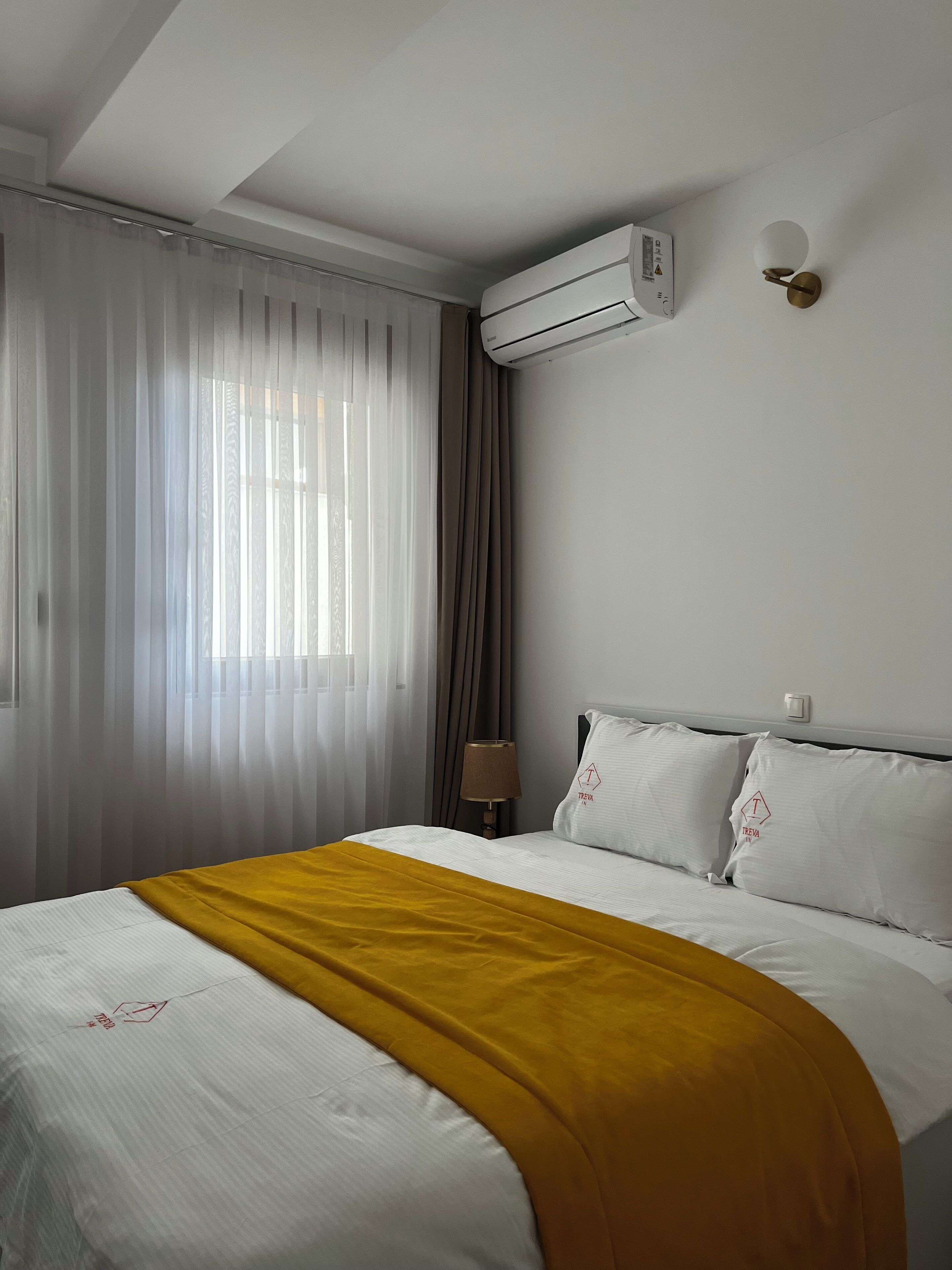 A hotel room with a bed, a bedside lamp, an air conditioning unit, and a window with curtains.