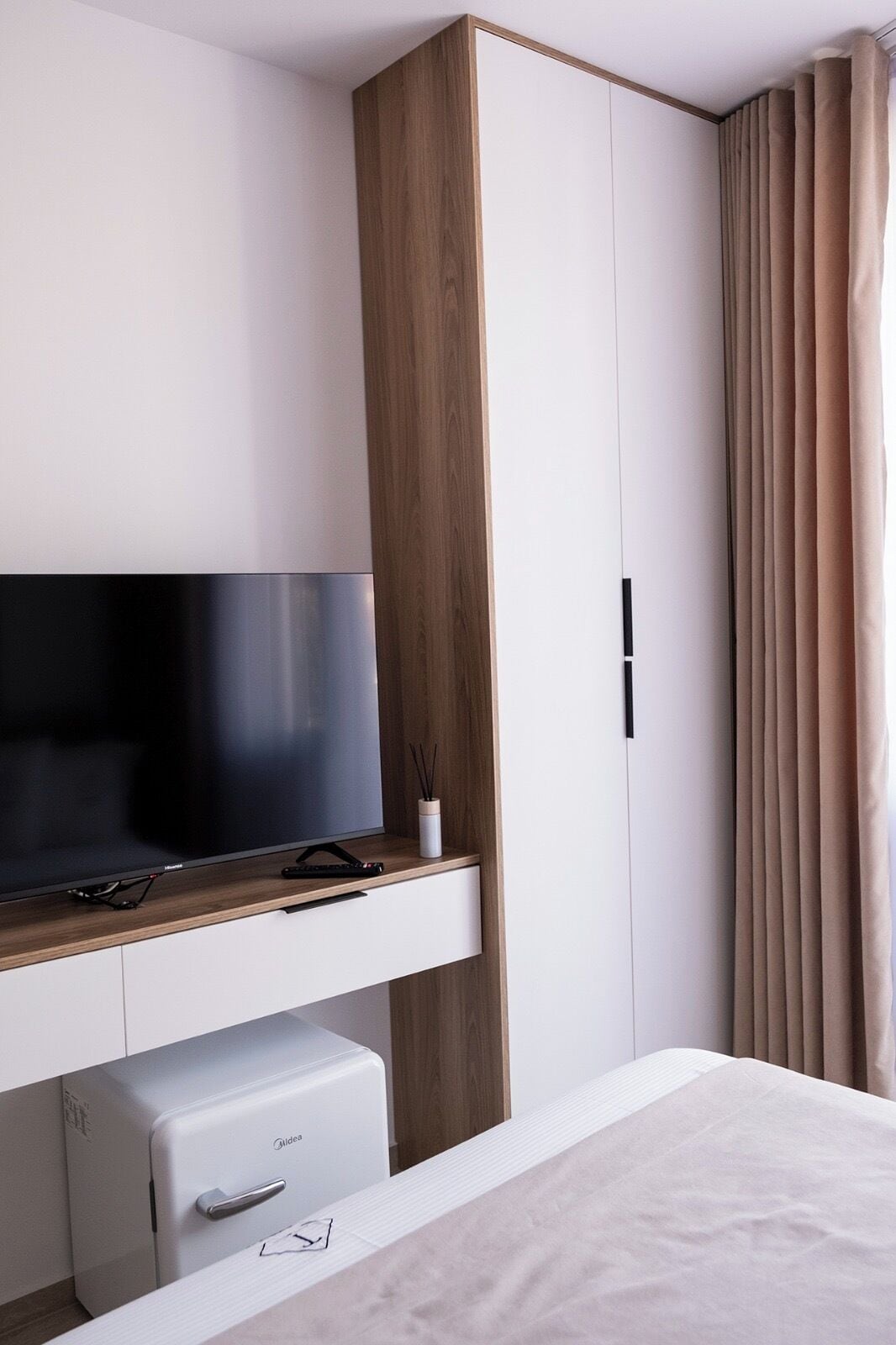 Ein modernes Hotelzimmer mit Flachbildfernseher auf einer Holzkonsole, einer Minibar und einem Kleiderschrank mit Spiegel.