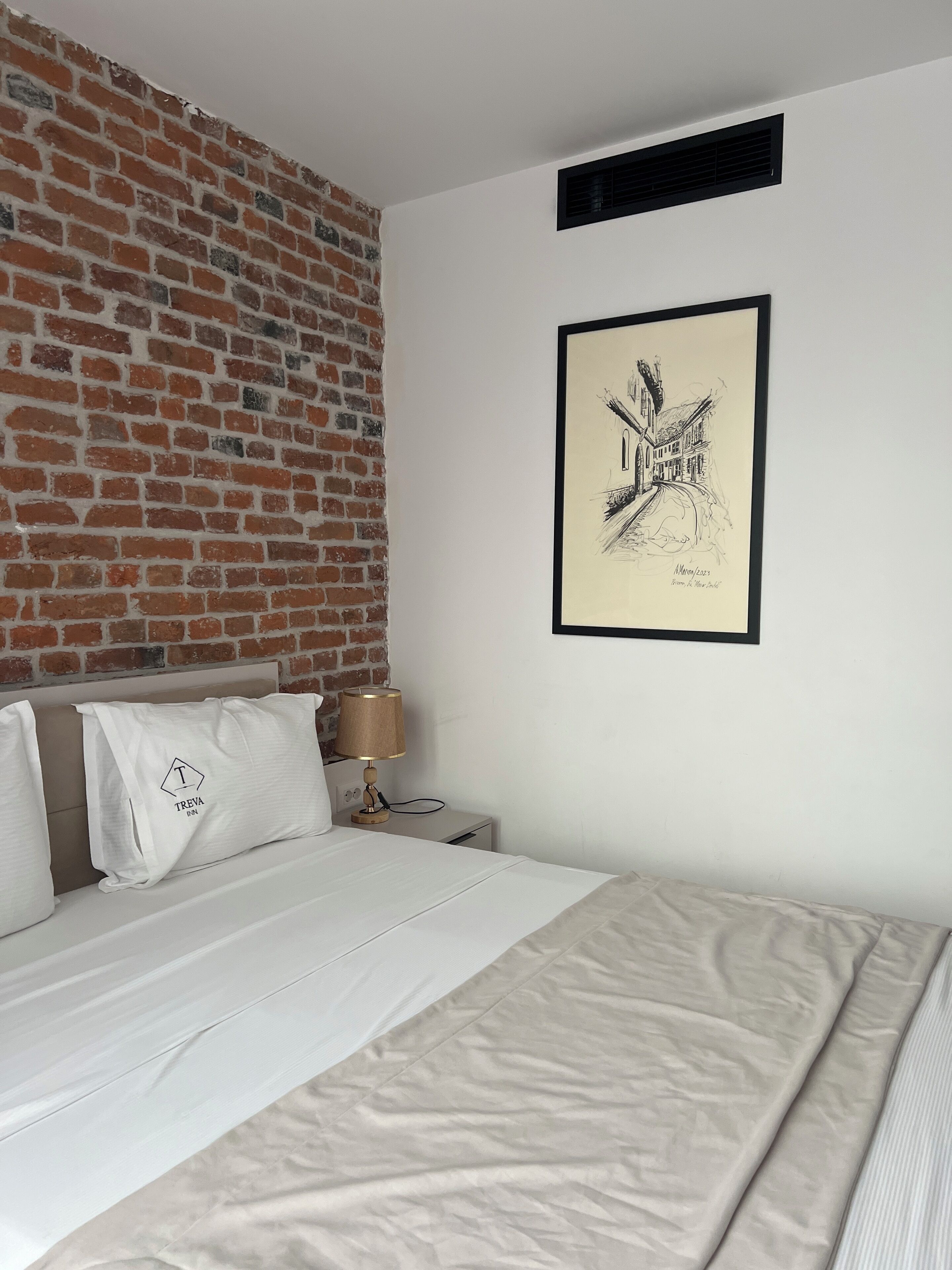 A hotel room with a brick wall, a framed artwork, a bed with white bedding, and a nightstand with a lamp.