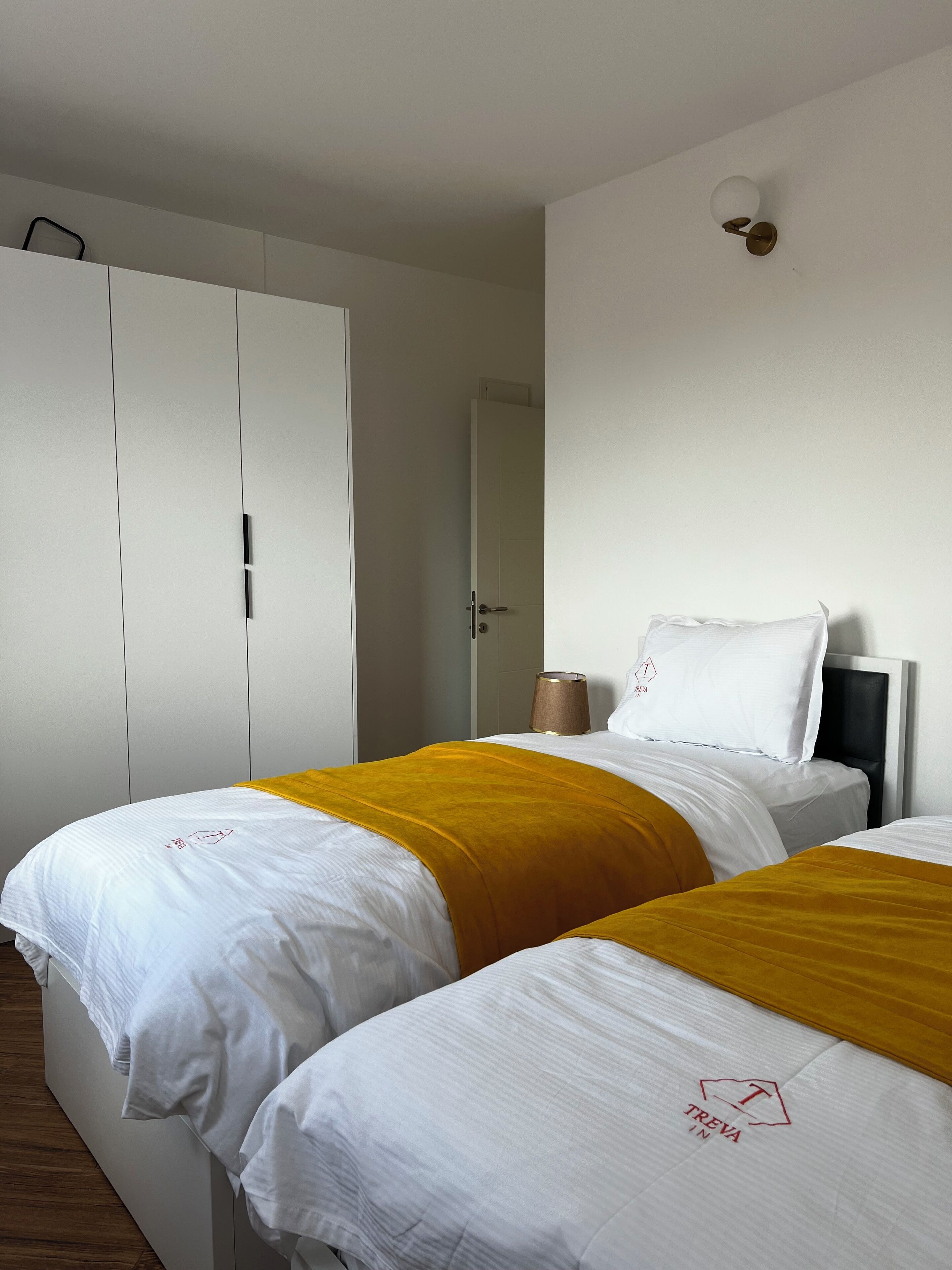 Et hotelværelse med to senge, hvidt sengetøj, et gult pled og et hvidt klædeskab samt en vægmonteret lampe.