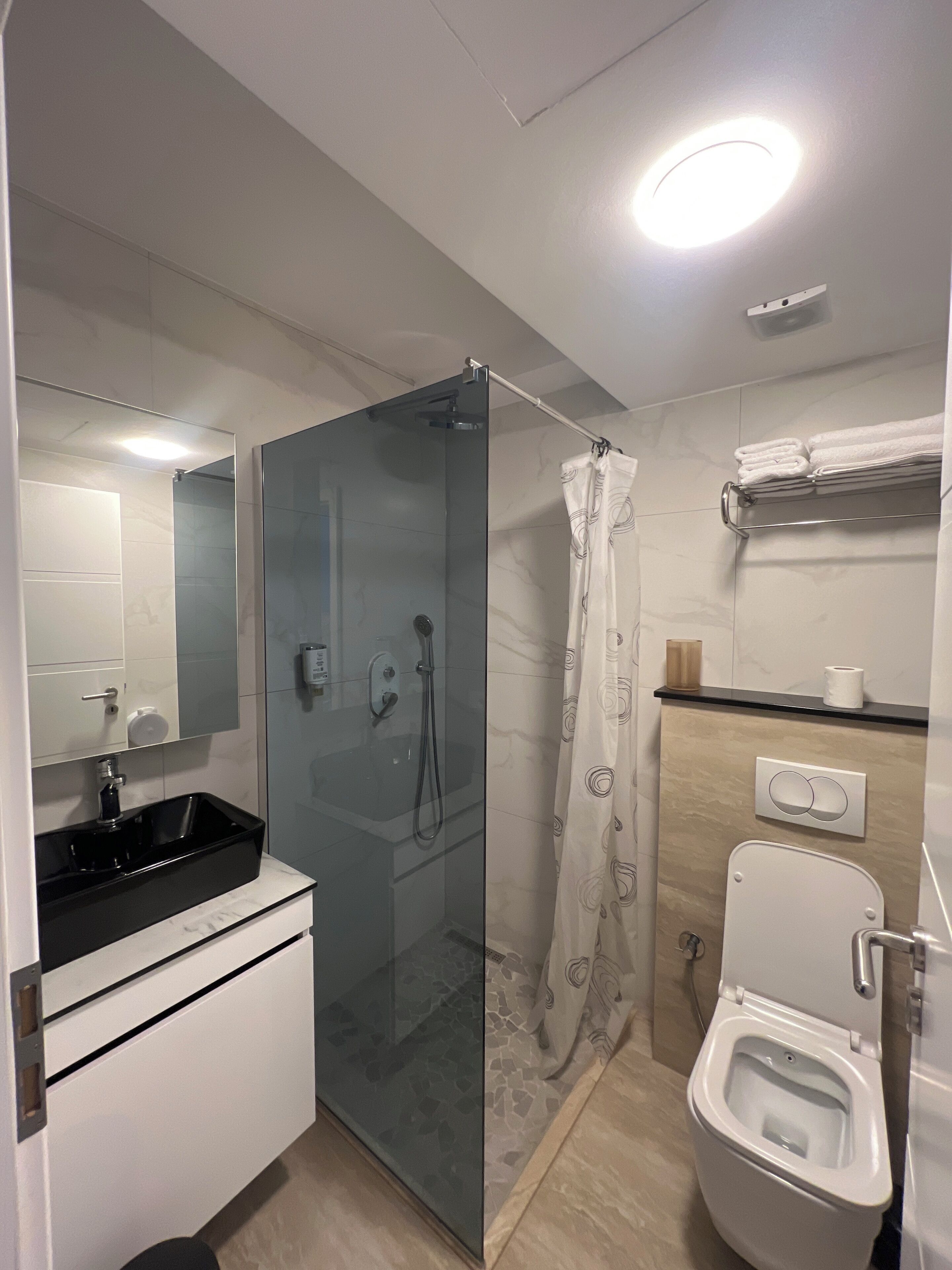 Un baño moderno con ducha envuelta en vidrio, inodoro blanco y lavamanos negro.