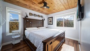 Un dormitorio con techo de madera, una cama con colcha estampada, una mesita de noche y un televisor montado en la pared, además de una ventana con vistas a árboles y montañas.
