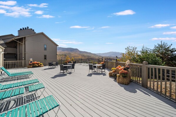 Una terraza de madera con mobiliario exterior, vistas a montañas y un edificio con chimenea.