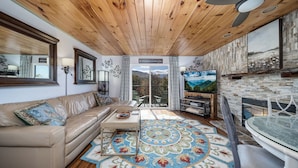 Un salón acogedor con techo de madera, televisor grande, chimenea y vistas al exterior.