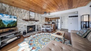 Una sala de estar moderna con chimenea, techo de madera, pared de piedra y un televisor grande.