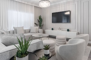 Una sala moderna con sofá, pufs, una mesita de centro, televisor y un mueble bajo en color blanco.