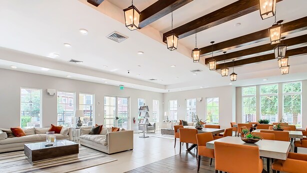 Eine großzügige Hotelhalle mit modernem Design, ausgestattet mit einer großen Sitzecke mit orangefarbenen Stühlen und einem Esstisch, einer Sofagarnitur und großen Fenstern, die für natürliches Licht sorgen.