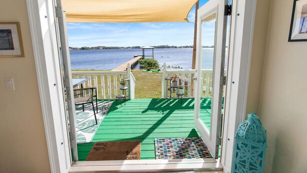 A view from a room with a sliding glass door to a deck with a green floor, a white railing, and a table with chairs. - LakeFront Sunrise Cottage (Sebring)