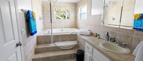 Un baño con bañera, lavabo y ducha. Hay escalones que dan acceso a la bañera, y una ventana que proporciona luz natural.