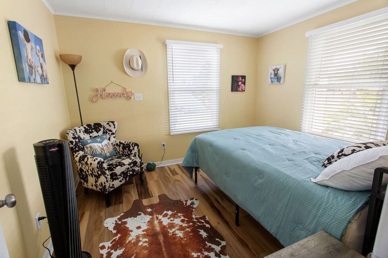 A bedroom with a cowhide rug, a cow print chair, a bed with a blue bedspread, and a wall-mounted hat.