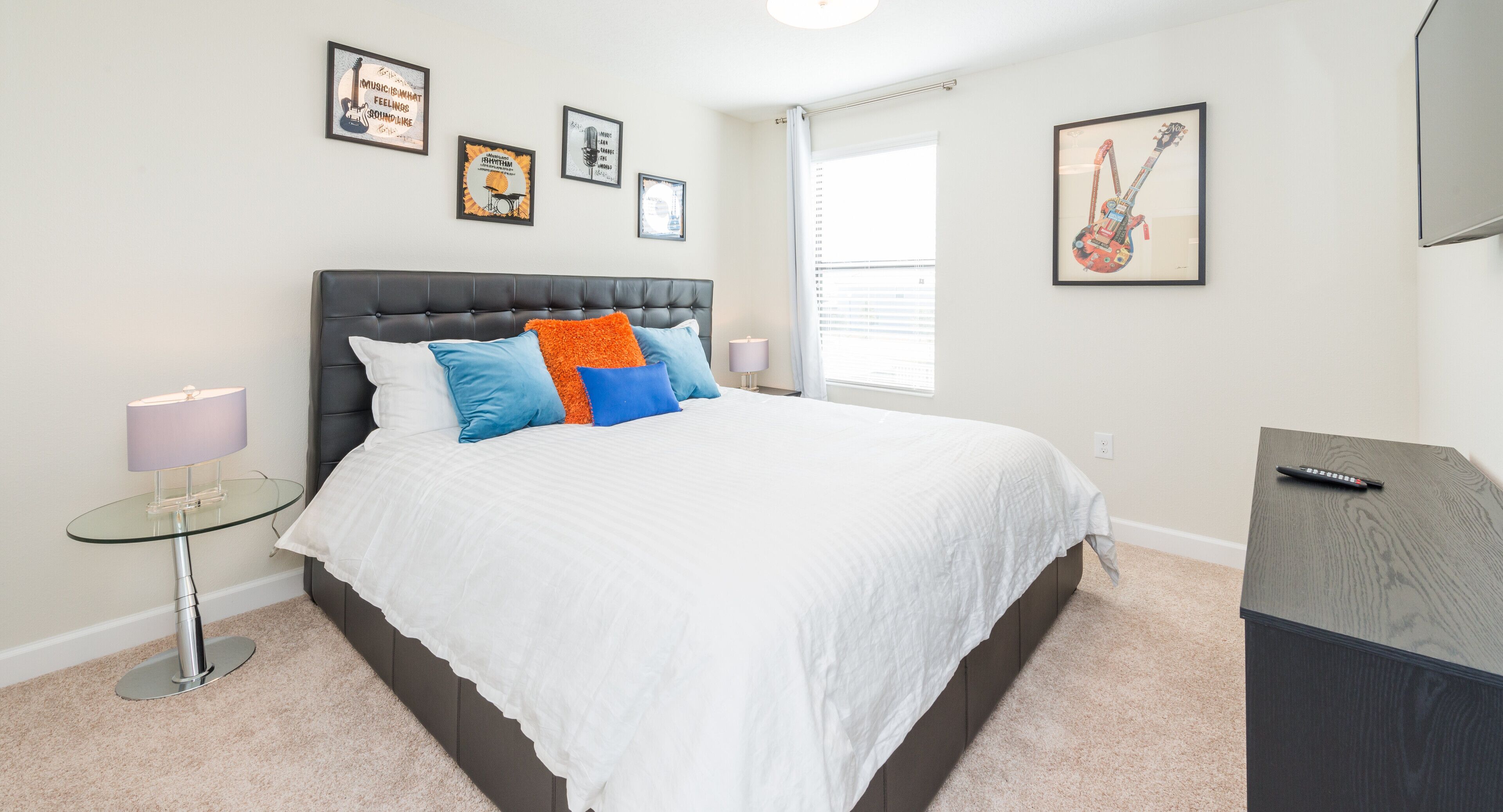 A bedroom with a bed, a small table with a lamp, a dresser, and framed pictures on the wall.