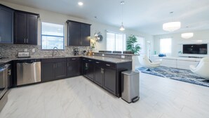 A modern kitchen with dark cabinets, a dishwasher, and a built-in oven. There is a dining area with a white sofa and a TV.
