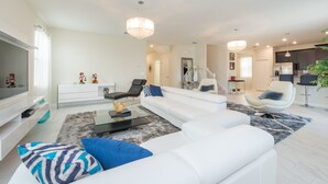 A modern living room with a white sofa, a glass coffee table, and a TV. There is a staircase leading to another level.