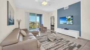 A modern living room with a beige sofa, a glass coffee table, a TV on the wall, and a balcony with a view. - Last Minute Disney Getaway! Themed Bedrooms & Private Balcony. Book Now! #113577 (Orlando)