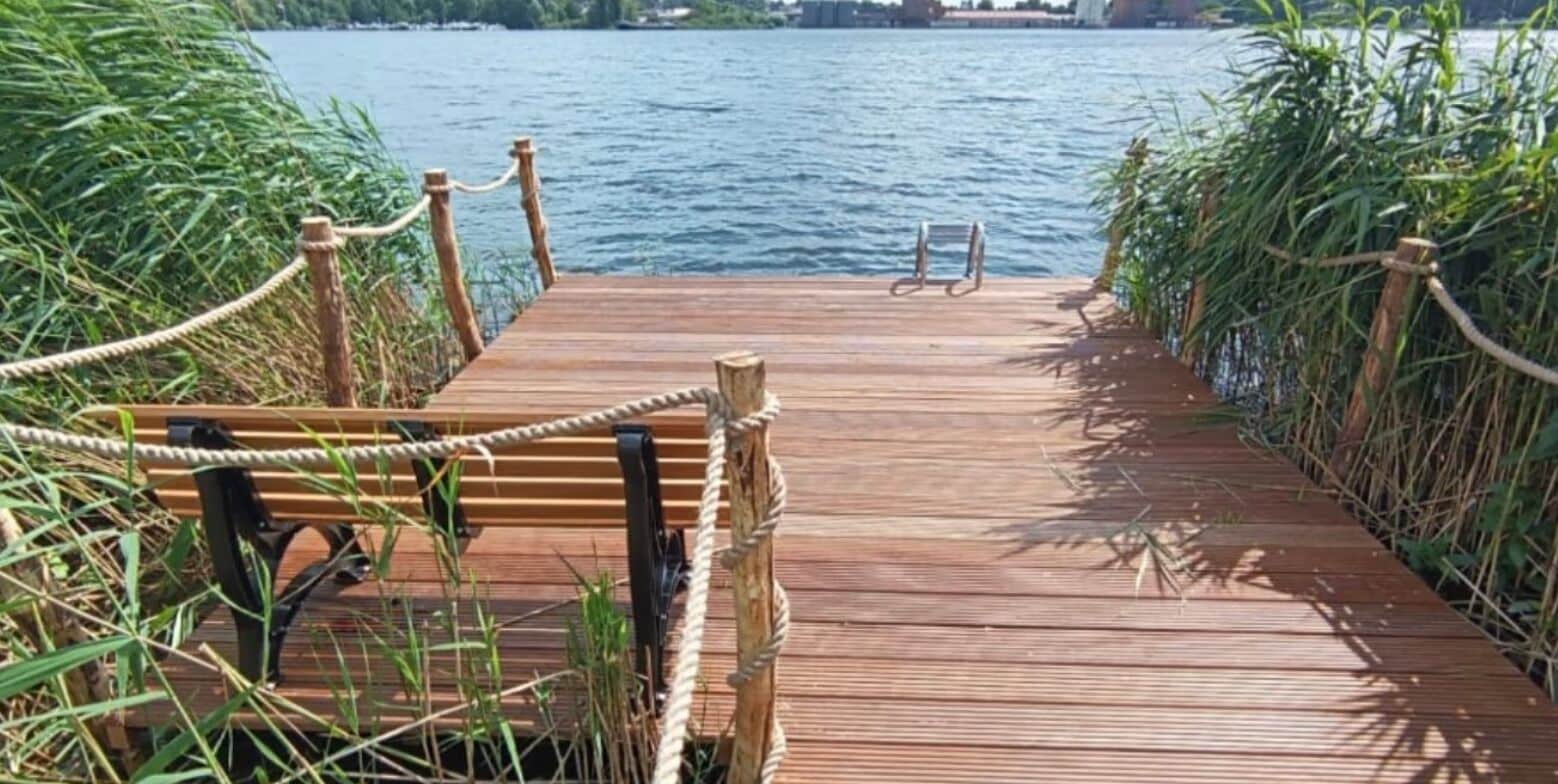 Een houten looppad dat leidt naar een bank aan het water, omringd door hoge gras.'