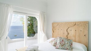 A bedroom with a large window, a wooden headboard, a bed with white linens, and a view of the sea.