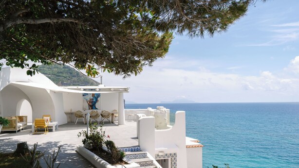 A coastal villa with a terrace overlooking the sea, furnished with white sofas and chairs, and a dining area with a table and chairs.