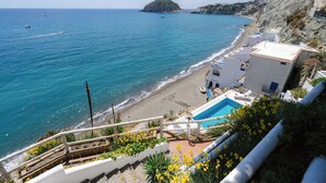 A coastal view with a beach, a swimming pool, and a hillside with buildings.
