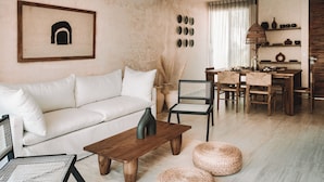 Een woonkamer met een witte bank, een houten salontafel, rieten stoelen en een eethoek met een houten tafel en stoelen.
