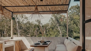 Een terras buiten met rieten dak, een eettafel gedekt voor twee personen en een zithoek met kussens.