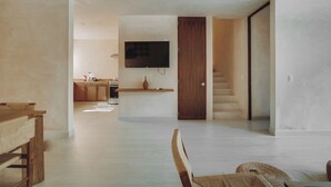 Een ruime woonkamer met een houten eettafel, een bank en een aan de muur bevestigde televisie. In de achtergrond is een keuken te zien met houten keukenkastjes en een aanrecht.