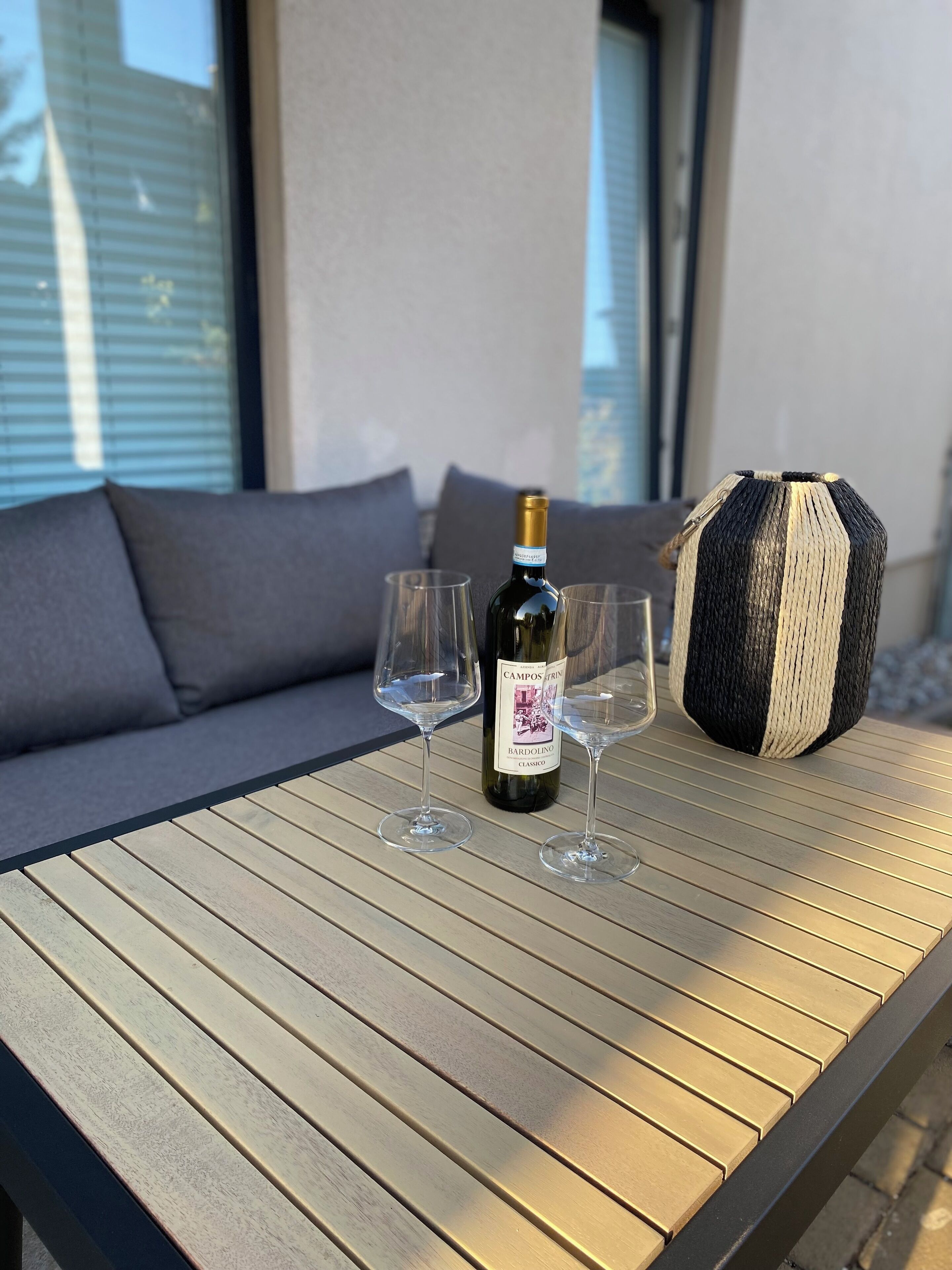 Une table en bois, sur laquelle se trouvent une bouteille de vin, deux verres et un panier en osier, dans un espace aménagé en plein air.