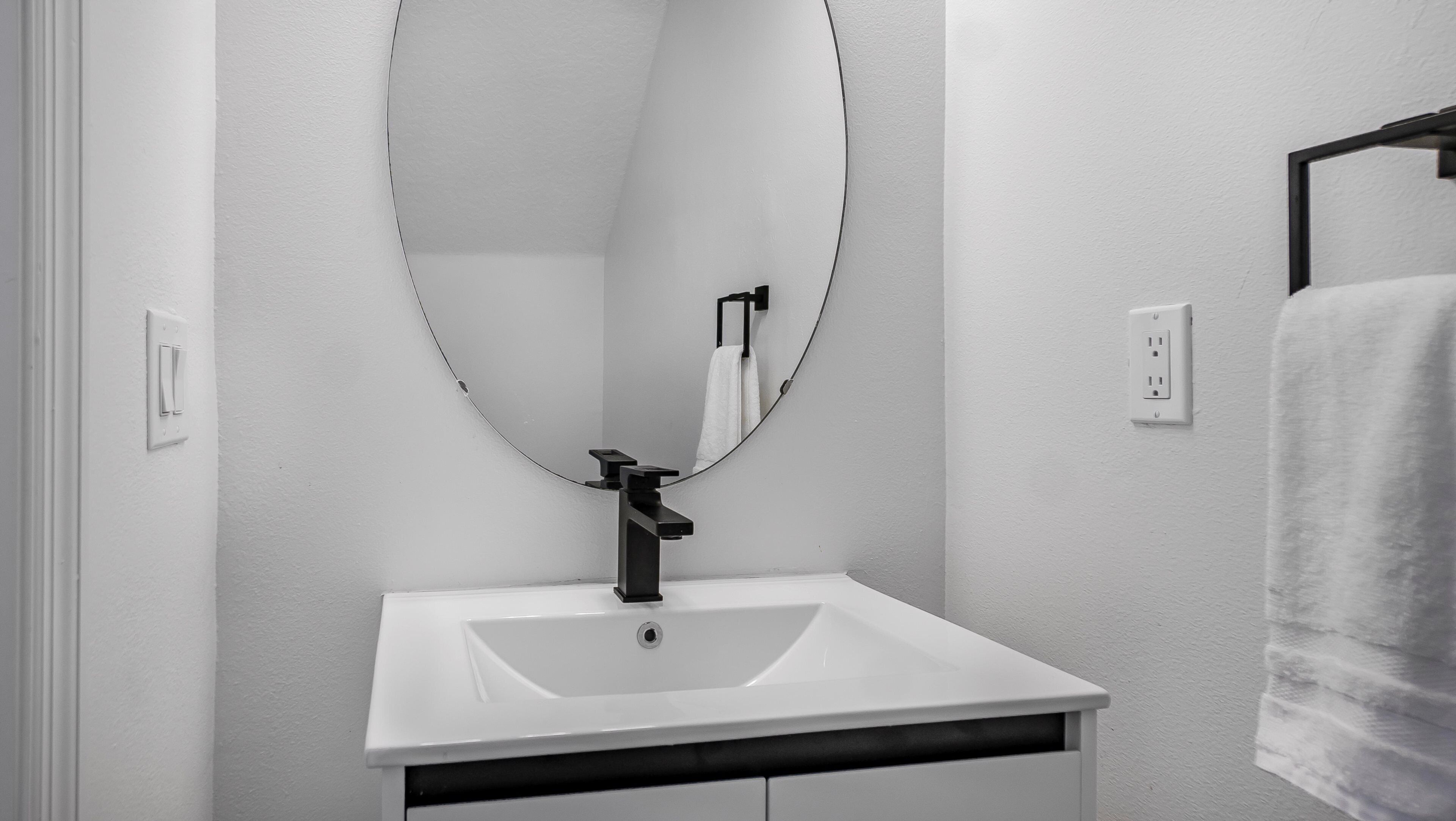 Une salle de bain avec un lavabo blanc, un robinet noir, un grand miroir ovale et une serviette suspendue à un porte-serviettes noir.