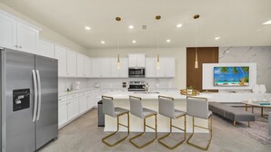 A modern kitchen with white cabinets, a stainless steel refrigerator, a central island with bar stools, a microwave, and a TV mounted on the wall.
