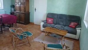 Ein Wohnzimmer mit einer Couch, zwei Sesseln, einem Holzschrank und einem Esstisch mit roter Tischdecke.