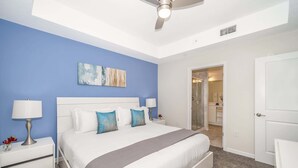 Une chambre à coucher comprenant un lit, deux tables de chevet, un ventilateur de plafond et une porte donnant sur une salle de bain.
