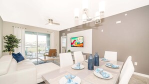 Un espace repas moderne avec une grande table dressée pour quatre personnes, des chaises blanches et un ventilateur de plafond.
