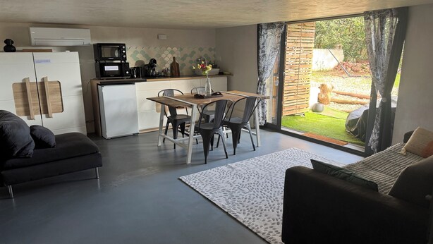 Ein modernes Wohnzimmer mit einer Couch, einem Esstisch mit Stühlen, einer Küchenzeile inklusive Kühlschrank, Mikrowelle und Kaffeemaschine. Ein Schiebetürenfenster führt zu einem Garten.