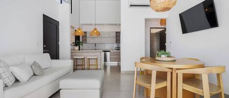 A modern living room with a white sofa, a dining area with a wooden table and chairs, and a kitchen in the background.