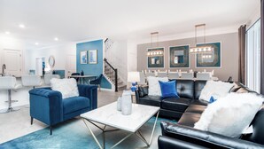 Una sala moderna con un sofá azul, un sofá de cuero negro y una mesa de centro blanca. Se ve una escalera que conduce a otro nivel, y un comedor con una araña.