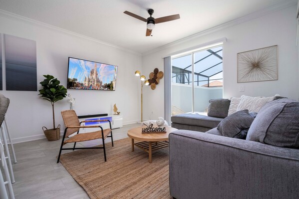 Ein modernes Wohnzimmer mit einem grauen Sofa, einem Holztisch, einem Korbstuhl, einem Fernseher und einem großen Fenster mit Blick.