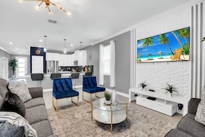 A modern living room with a large TV, two blue armchairs, a glass coffee table, and a kitchen in the background.