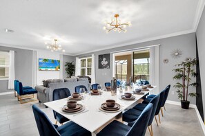 A dining area with a long table set for six, surrounded by blue chairs, with a sofa and a TV in the background. - Last Minute Disney! Private Pool, Game Room & Themed Bedrooms. Book Now! #115455 (Orlando)